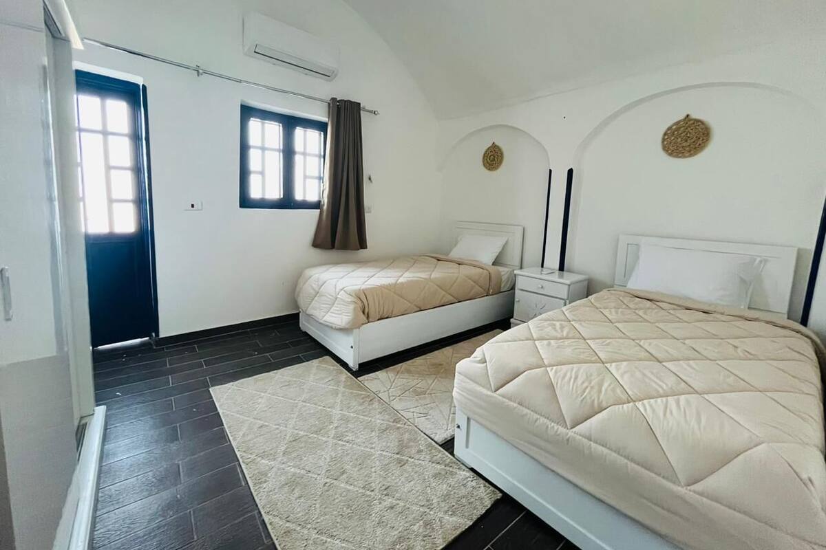 A bedroom with two beds, a window with curtains, a nightstand, and a wall-mounted air conditioner.