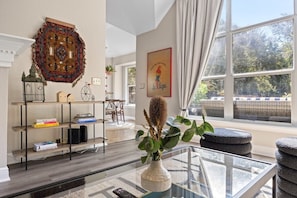 A living room with a glass coffee table, a bookshelf, a decorative rug, and a window with a view of trees.