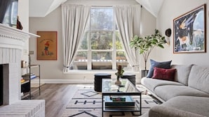 A living room with a fireplace, a sofa, a coffee table, and a large window with curtains. - Eclipse · Artist Retreat @ Cypress Creek | Hotub | Sleeps 9 (Cedar Park)