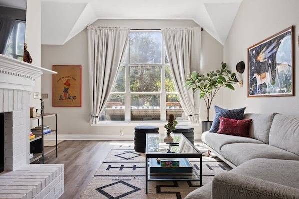 A living room with a fireplace, a sofa, a coffee table, and a large window with curtains.