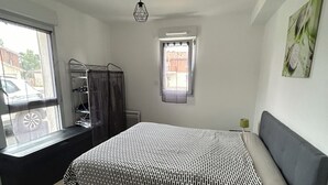 Un dormitorio con cama, ventana, radiador, estante y vistas al exterior.