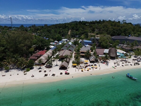 An aerial view of a beach resort with thatched-roof buildings, a sandy beach, and clear turquoise waters.
