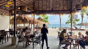 Un espace de restauration en plein air, avec des tables et des chaises, un toit de chaume et une vue sur la plage et l’océan.