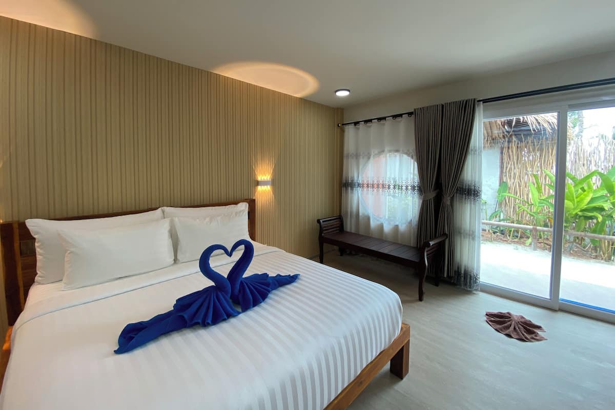 A hotel room with a bed, a bench, a window with curtains, and a view of greenery outside.