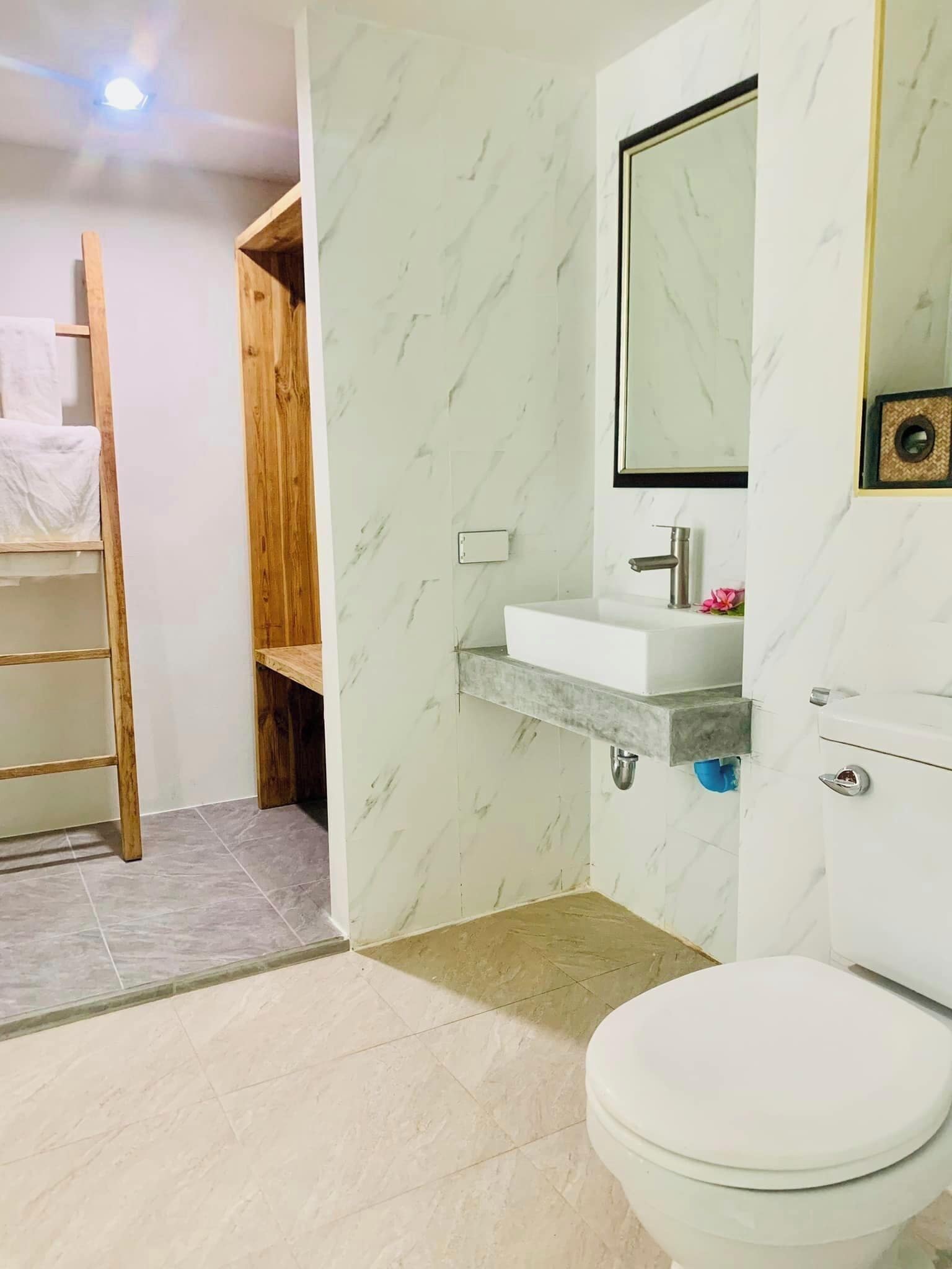 A bathroom with a white toilet, a wooden shelf, a marble-patterned wall, and a white sink with a mirror above it.