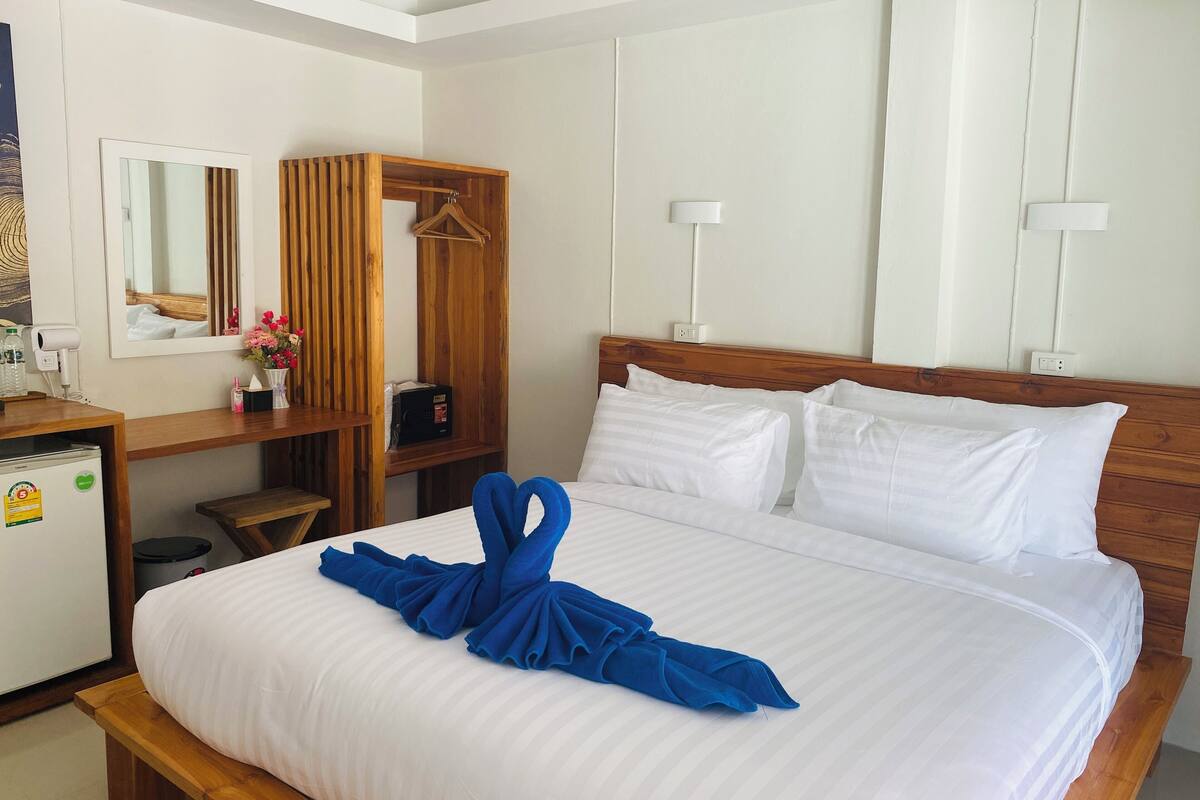A hotel room with a neatly made bed, a wooden bedside table, a mirror, a wooden wardrobe, and a small desk with a lamp.