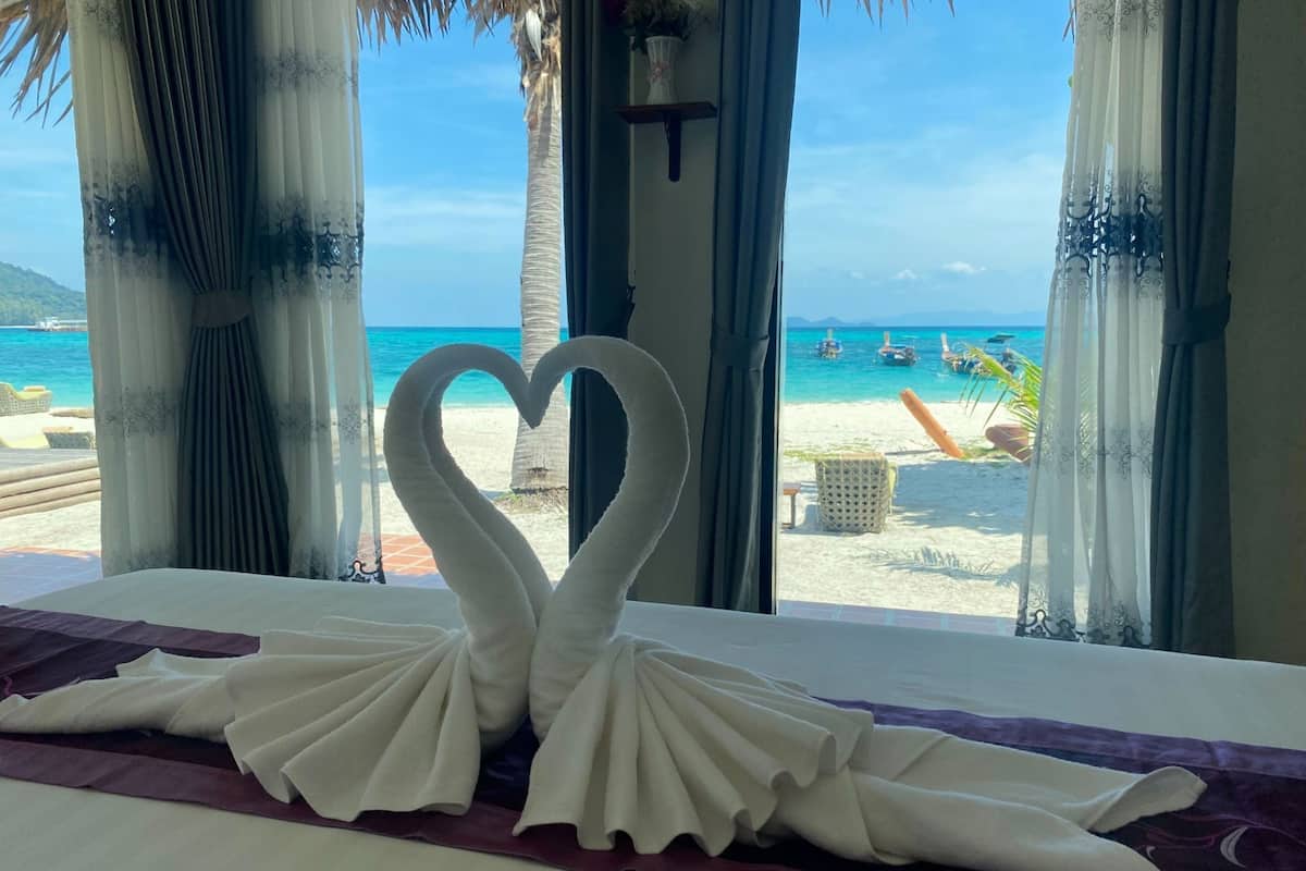 A bed with a heart-shaped towel arrangement and a view of a beach with boats.