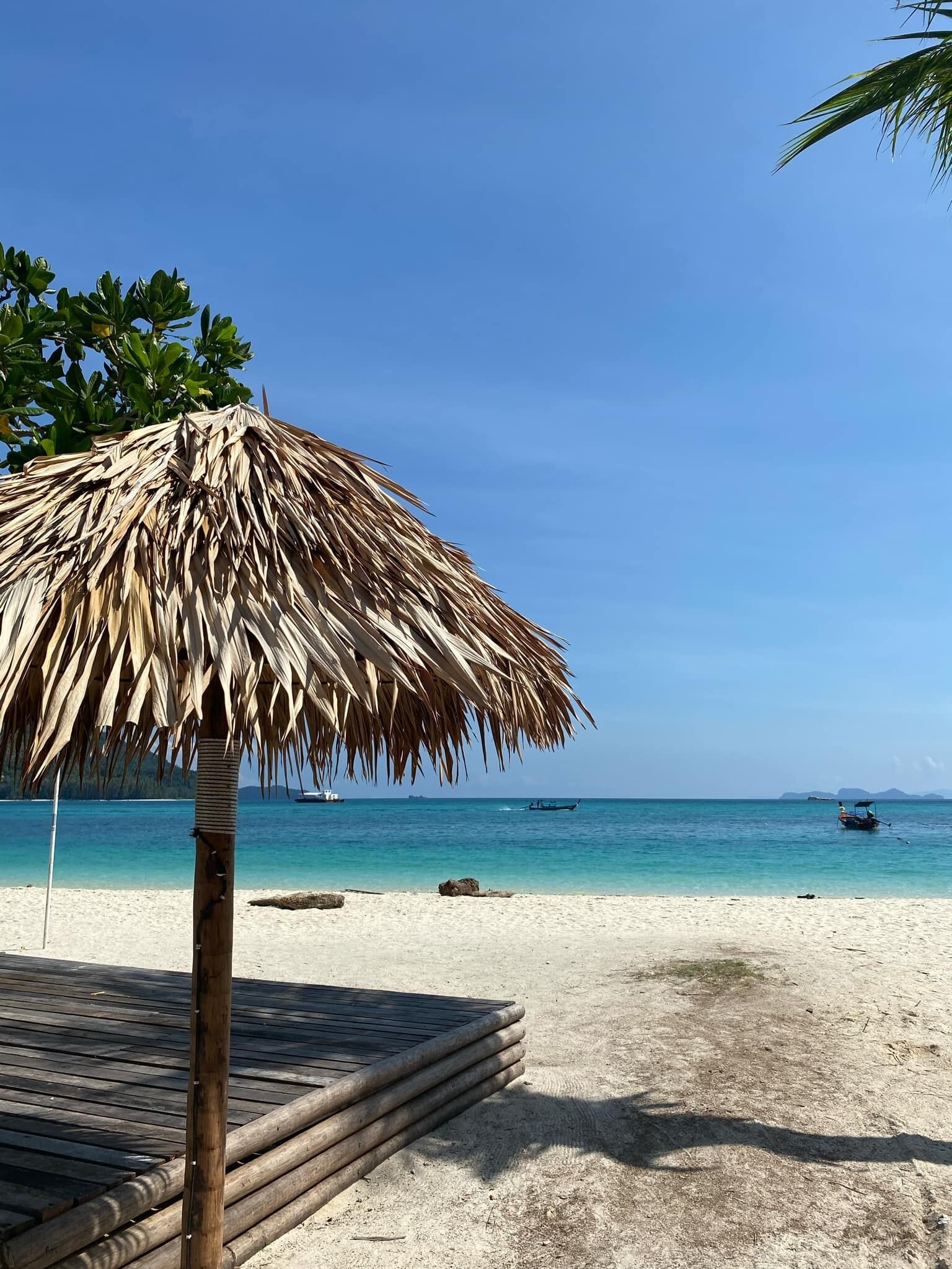 Una spiaggia con un ombrellone di paglia, acqua cristallina e barche in lontananza.
