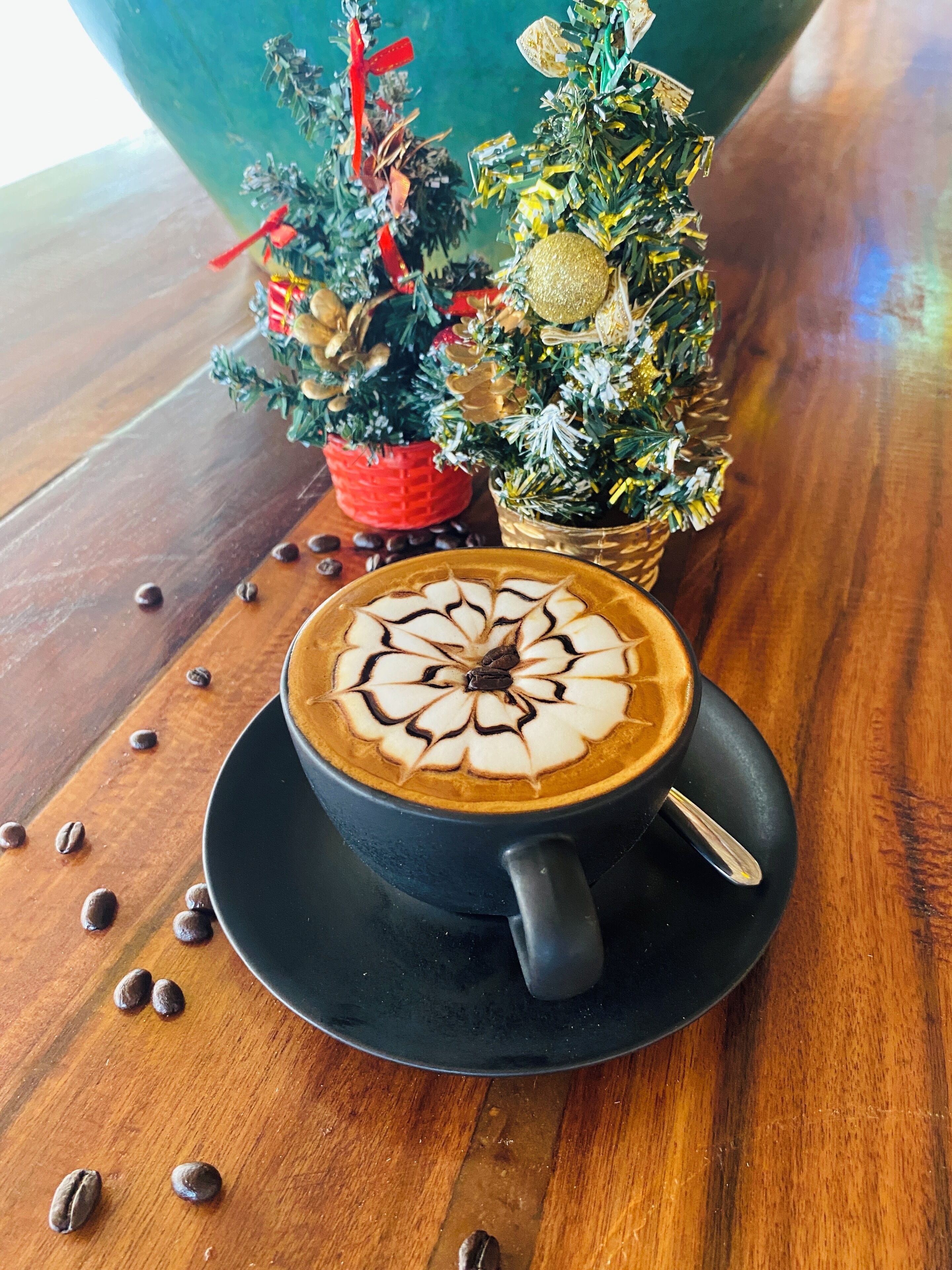 A cup of coffee with latte art on a wooden table, surrounded by coffee beans and small Christmas trees.