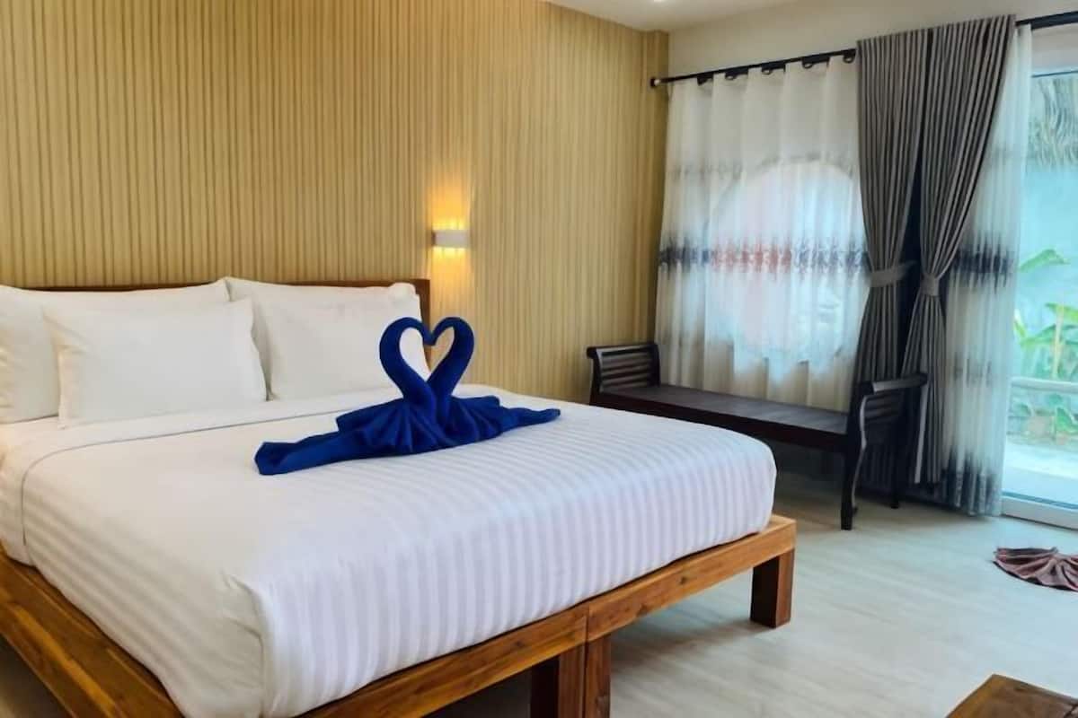 A hotel room with a bed, a wooden bench, a chair, a window with curtains, and a wall-mounted light.