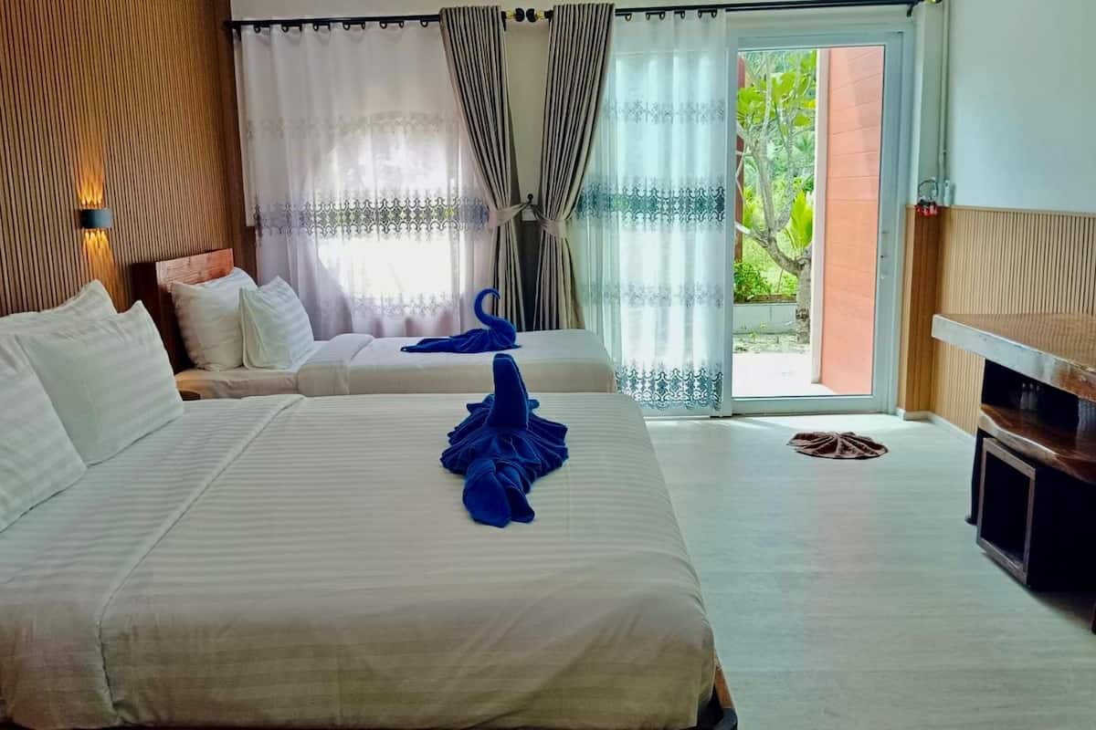 A hotel room with two beds, a wooden headboard, a small table, and a view of greenery through the window.