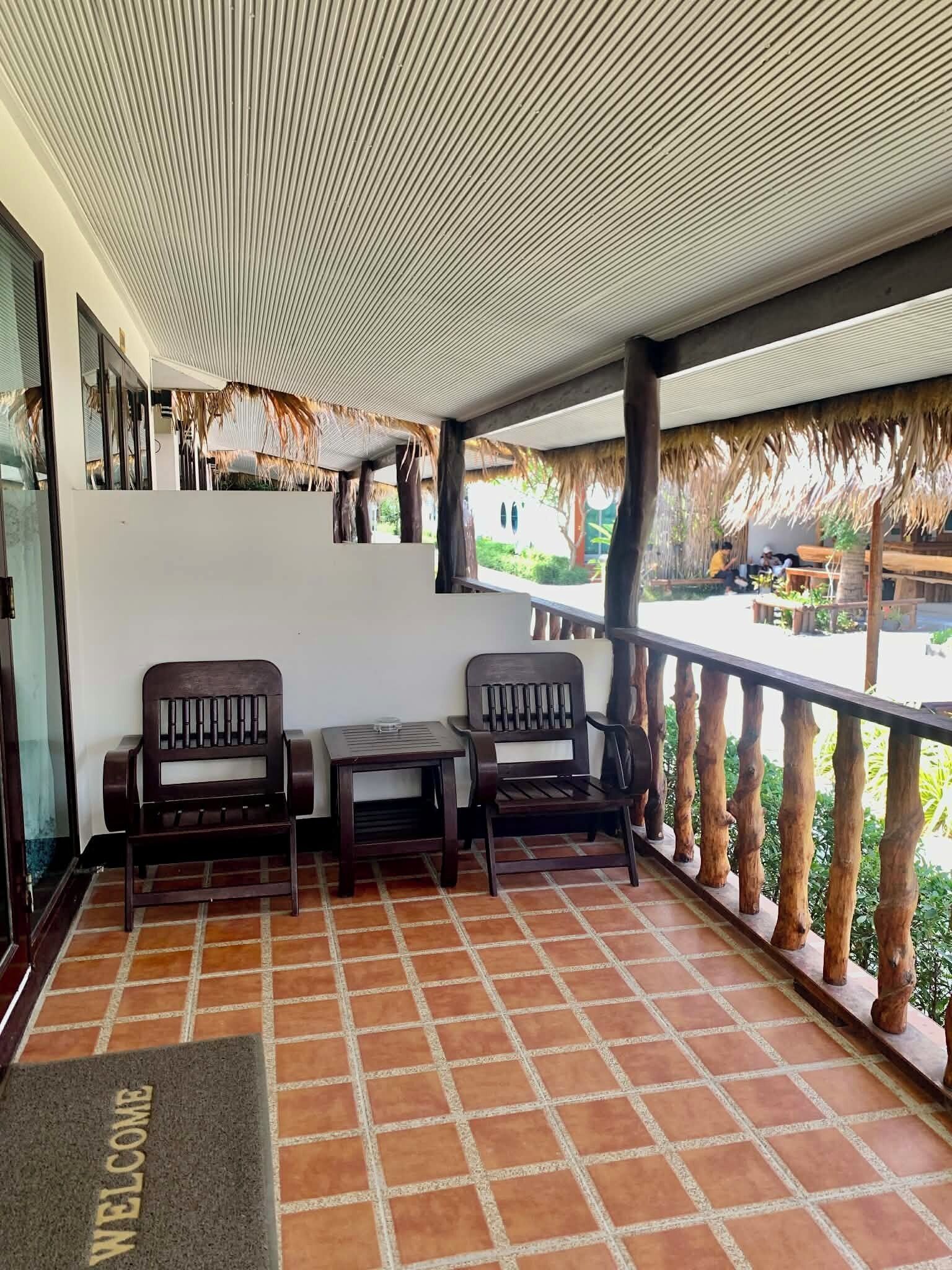A covered outdoor seating area with two wooden chairs and a small table, a welcome mat, and a view of a thatched-roof building and greenery.