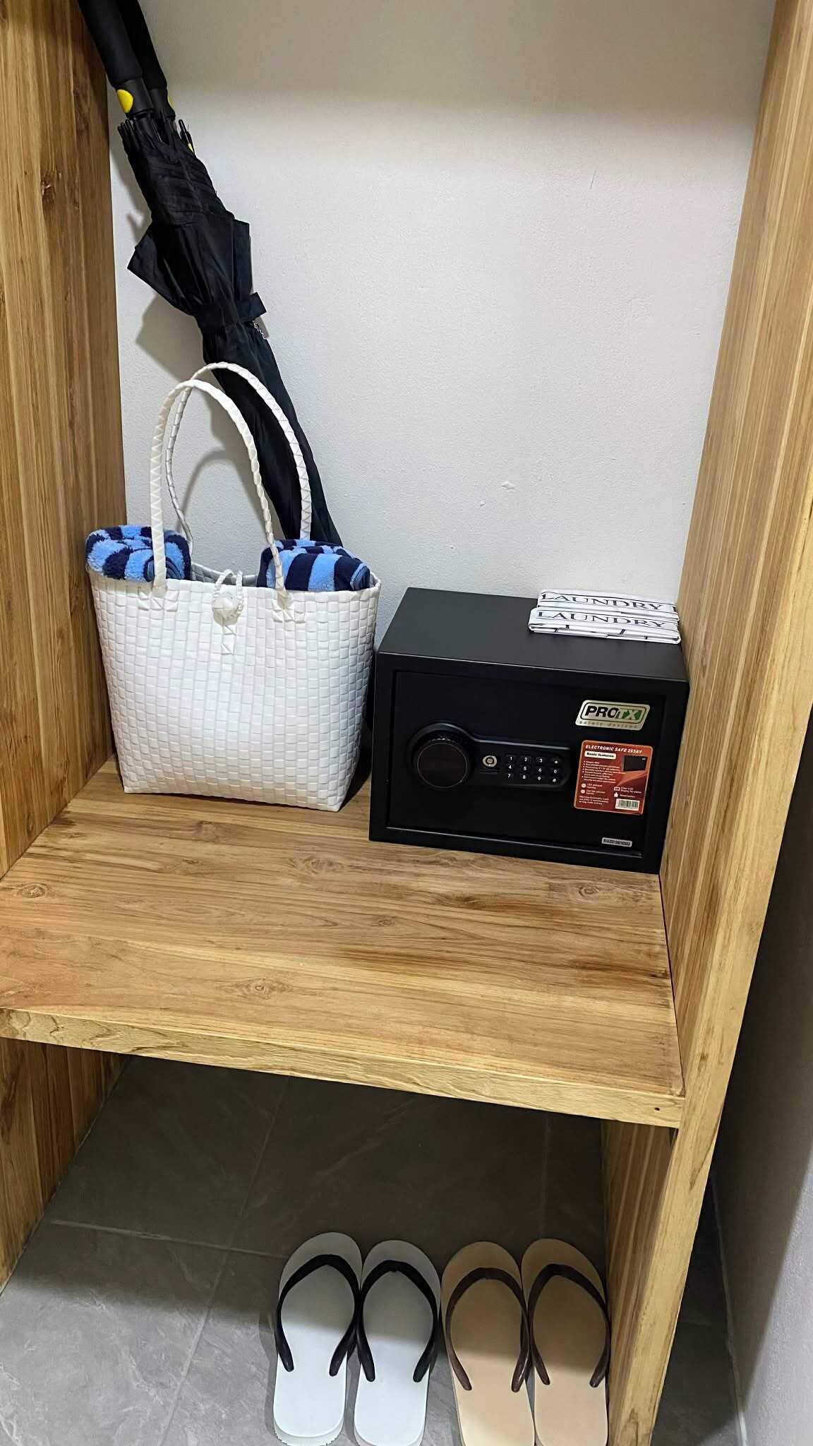A wooden shelf with a white quilted bag, a black electronic safe, and a pair of flip-flops.