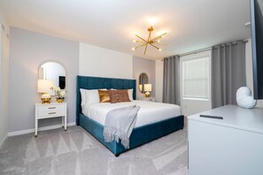 A modern bedroom with a blue upholstered bed, white nightstands, a desk, and a large mirror.