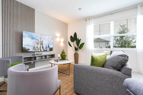 A modern living room with a grey sofa, a white armchair, a TV mounted on the wall, and a large window with blinds.