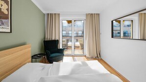 A hotel room with a bed, a chair, a small table, a window with curtains, and a view of buildings outside. - Flora Collections - Studio room with shared kitchen (Vienna)