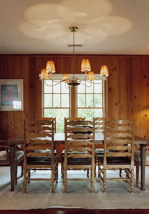 Un comedor con una mesa y sillas de madera, una lámpara de araña y un ventanal con un cartel.