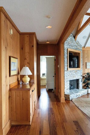 A wooden interior with a fireplace, a hallway leading to another room, and a lamp on a wooden cabinet.