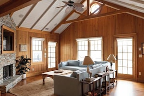 A spacious living room with wooden walls, a stone fireplace, a sofa, and a ceiling fan. - The Holiday | 5BR 5BA Coastal Retreat (Wrightsville Beach)
