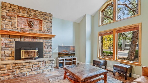 A living room with a stone fireplace, a television, and a large window with a view of trees. - Bluebird Song | Prime Flagstaff Location | Outdoor Retreat with Fenced Yard and Firepit (Flagstaff)