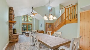 A spacious dining area with a large wooden table and white chairs, a stone fireplace, and a wooden staircase. - Bluebird Song | Prime Flagstaff Location | Outdoor Retreat with Fenced Yard and Firepit (Flagstaff)