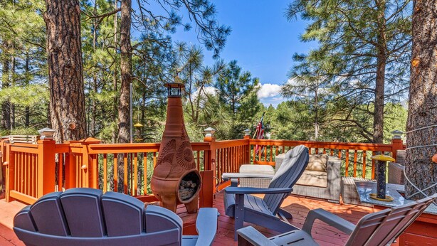 A deck with Adirondack chairs, a stone fireplace, and a view of pine trees. - ACE - A Hole in One! | Right on the Golf Course | Hot Tub |  Mt Views & Pines (Flagstaff)