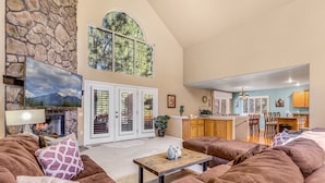 A spacious living room with a stone wall, a fireplace, and a large window overlooking a deck. - ACE - A Hole in One! | Right on the Golf Course | Hot Tub |  Mt Views & Pines (Flagstaff)