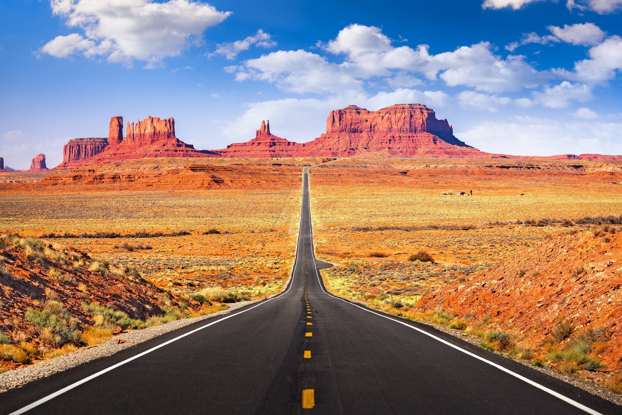 Eine gerade Straße, die zu einer gebirgigen Landschaft mit roten Felsformationen führt.