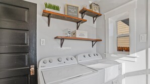 A laundry room with two Whirlpool washers, wooden shelves above holding various items, and a window with blinds.