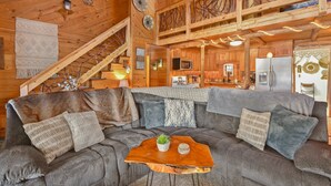 A cozy cabin interior with a large gray sofa, a wooden coffee table, and a kitchen area with wooden cabinets and a refrigerator.