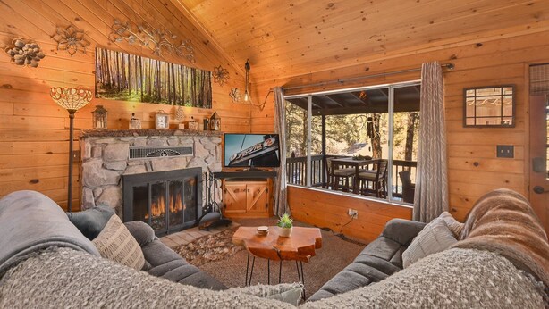 A cozy cabin interior with a stone fireplace, a television, and a large window overlooking a forest.