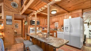 A wooden cabin interior with a kitchen featuring a granite countertop, bar stools, a refrigerator, and wooden cabinets.