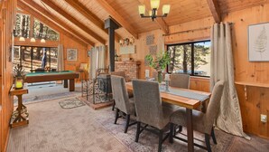 A cozy cabin interior with a pool table, dining table, and chairs, wooden beams, and large windows offering views of the forest.