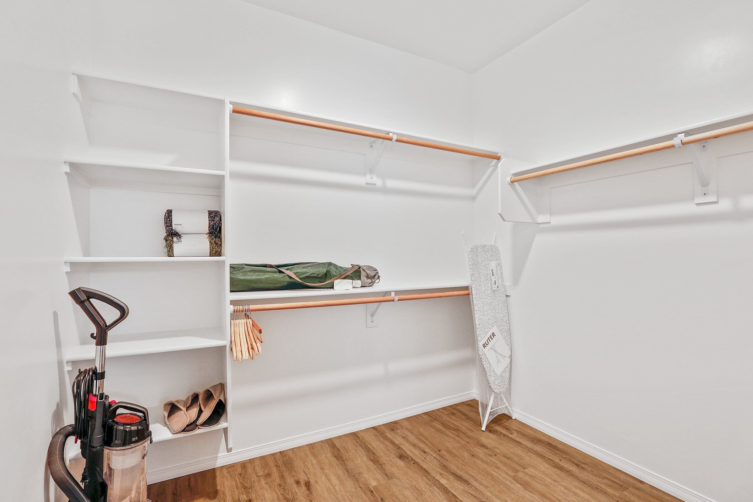 A walk-in closet with white shelves, a wooden floor, and a vacuum cleaner.