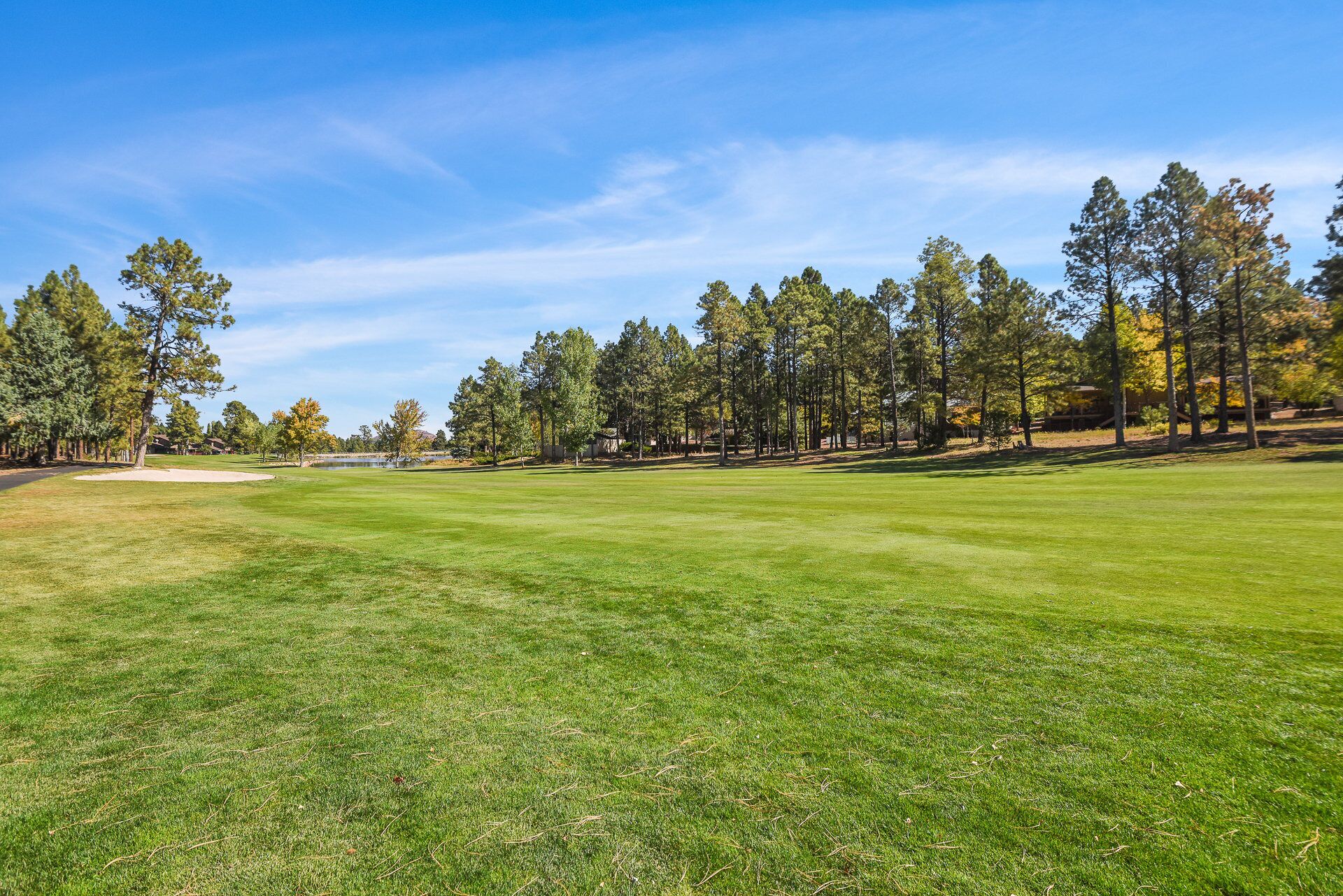 A well-maintained golf course with a clear sky, surrounded by tall trees and a few buildings in the background.