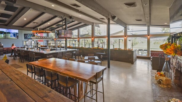 A spacious restaurant with a long wooden table and multiple seating areas, large windows offering views, and a bar area with a television screen.