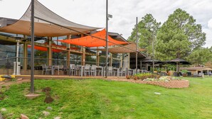 An outdoor seating area with tables and chairs, a building with large windows, and a grassy lawn with a flower bed.
