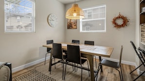 Un coin repas avec une table en bois, des chaises noires, un luminaire suspendu en fibres tressées, une horloge et une guirlande décorative accrochée au mur.