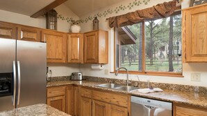 A kitchen with wooden cabinets, a stainless steel refrigerator, a Bosch dishwasher, and a window with a view of trees. - Burning Tree Lodge - Forest Views, Fire Pit, AC | Pet and Family Friendly (Flagstaff)