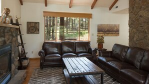 A cozy living room with a fireplace, leather sofas, and a large window overlooking a forest. - Burning Tree Lodge - Forest Views, Fire Pit, AC | Pet and Family Friendly (Flagstaff)