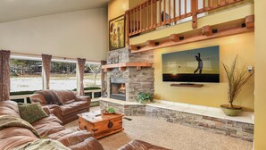 Amplia sala de estar con un sofá de cuero grande, una mesa de centro de madera y una chimenea de piedra. Un televisor está montado en la pared, y hay un balcón con barandilla de madera.