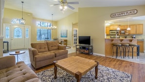 A living room with a sofa, a coffee table, and a TV. There's a kitchen area with wooden cabinets and a bar stool.