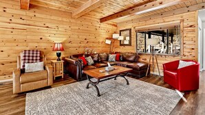A log cabin interior with a leather sofa, a red chair, a wooden coffee table, and a large window offering a view of trees.