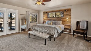A bedroom with a large bed, a ceiling fan, a wooden accent wall, and a view through a sliding glass door.