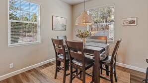 Un comedor con una mesa y sillas de madera, una lámpara colgante y dos ventanas con persianas.