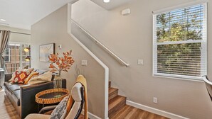 Una escalera que lleva a un piso superior, una ventana con persianas, un sofá con cojines estampados y un jarrón decorativo con hojas.