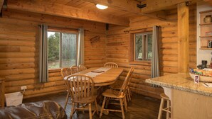 Interior de una cabaña de madera con una mesa de comedor, sillas, una barra y un ventanal con cortinas.