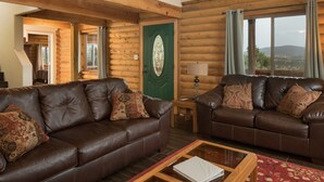 Interior de una cabaña de troncos con dos sofás de cuero, una mesa de centro de madera y una puerta verde con un panel de vidrio decorativo.