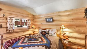 A log cabin bedroom with a bed, two bedside lamps, a window with curtains, and a picture on the wall.