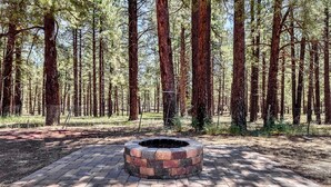 A circular brick fire pit surrounded by a paved walkway in a forested area. - Rustic Star | Authentic Log Cabin in the Woods | Near the Grand Canyon, Flagstaff & Williams (Bellemont)